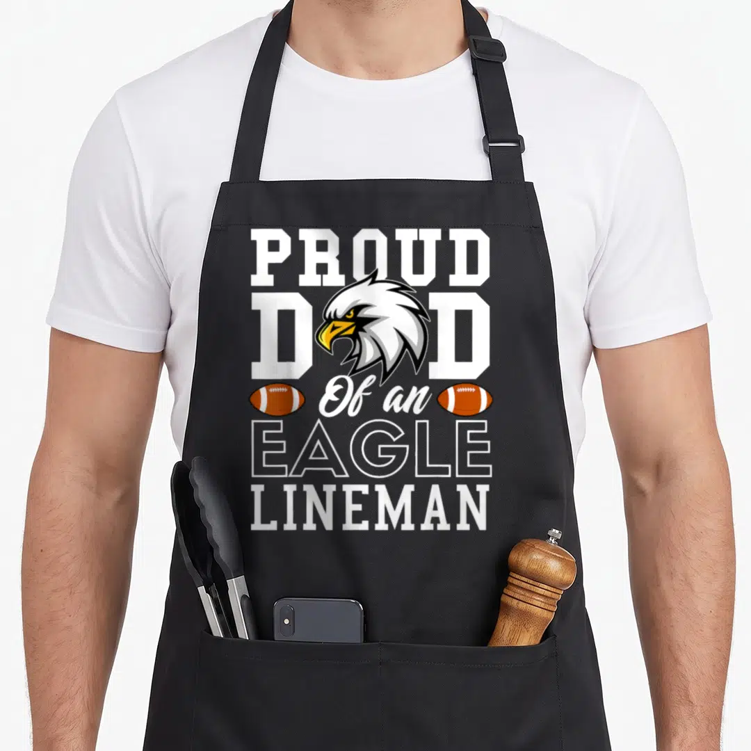 Proud eagles apron - Cartoon eagle head over white text PROUD DAD Of an EAGLE LINEMAN with two brown footballs — shown on a black cotton canvas apron against a white background