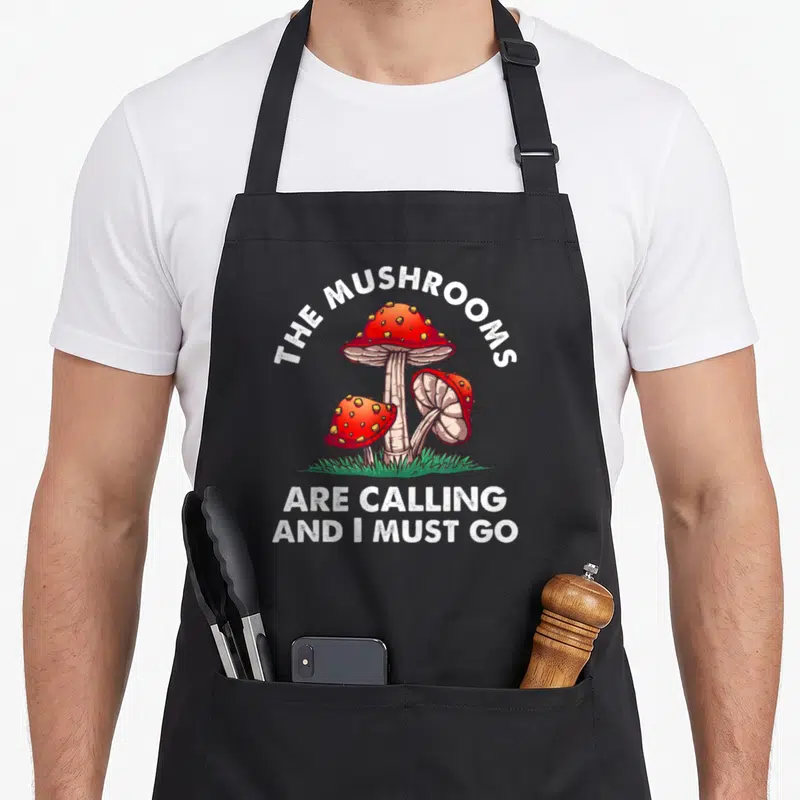 Funny calling mushroom apron - Three red spotted mushrooms in green grass with white text reading THE MUSHROOMS ARE — shown on a black cotton canvas apron against a white background