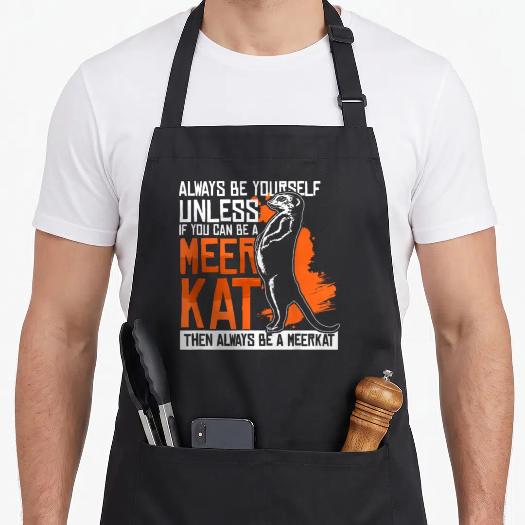 Vintage meerkat apron - Standing meerkat line art with orange splash and stacked text Always Be Yourself Unless You Can — shown on a black cotton canvas apron against a white background