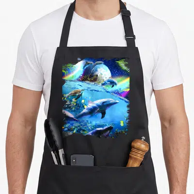 Galaxy dolphin apron - Surreal dolphins and turtle swimming near a planet with rainbows and palm leaves in blue, green — shown on a black cotton canvas apron against a white background