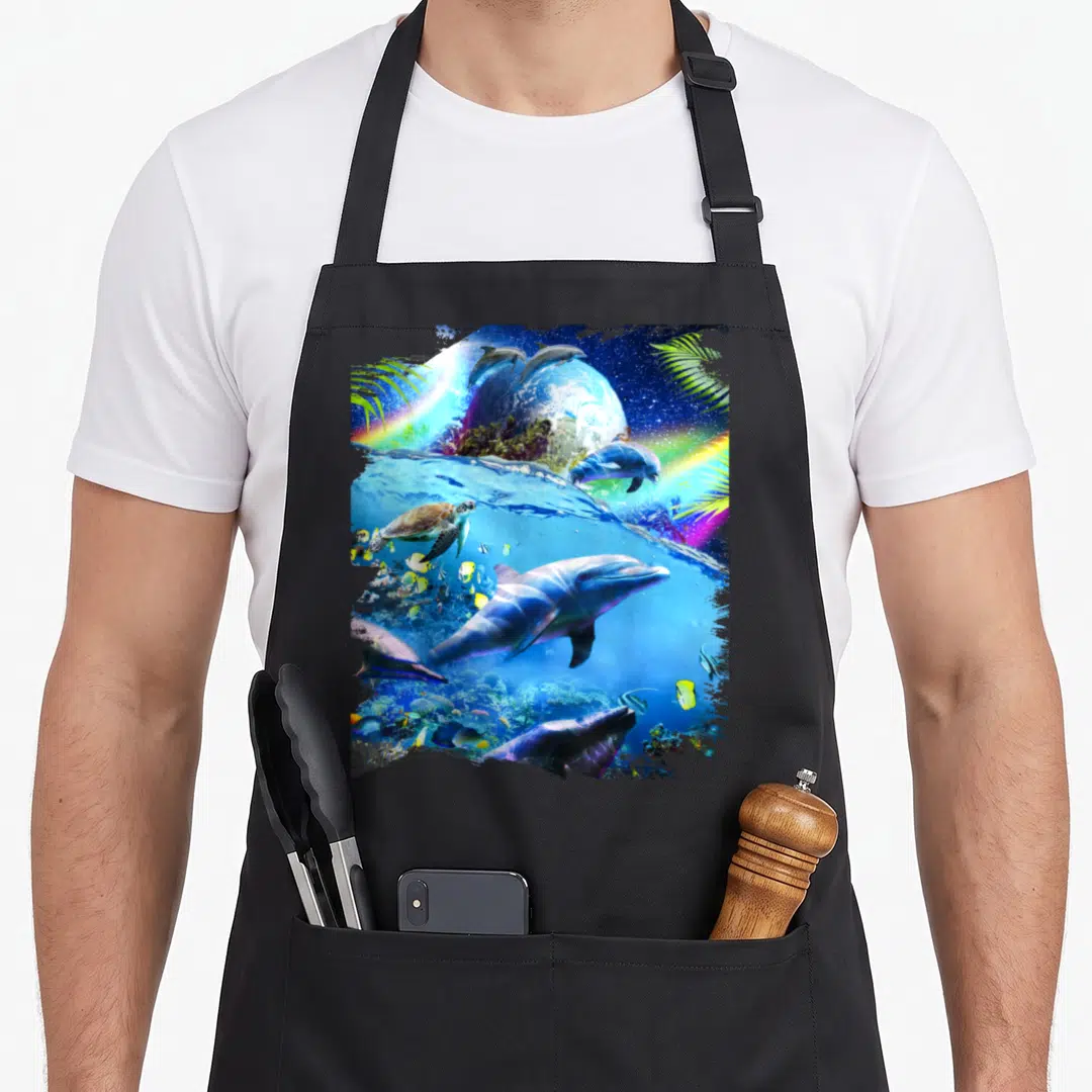 Galaxy dolphin apron - Surreal dolphins and turtle swimming near a planet with rainbows and palm leaves in blue, green — shown on a black cotton canvas apron against a white background