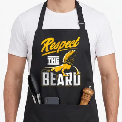 Respect beard apron - Yellow bearded dragon illustration with Respect THE BEARD text in white and yellow blocks — shown on a black cotton canvas apron against a white background