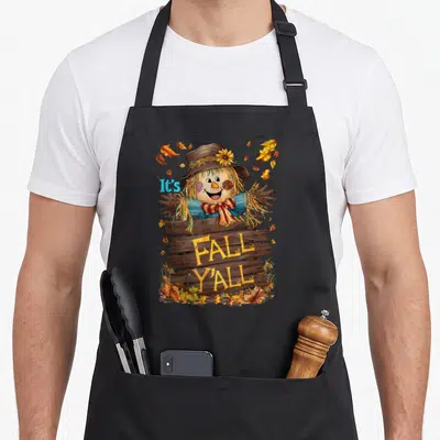 Fall yall thanksgiving apron - Cartoon scarecrow behind wooden sign reading It's FALL Y'ALL surrounded by brown yellow — shown on a black cotton canvas apron against a white background