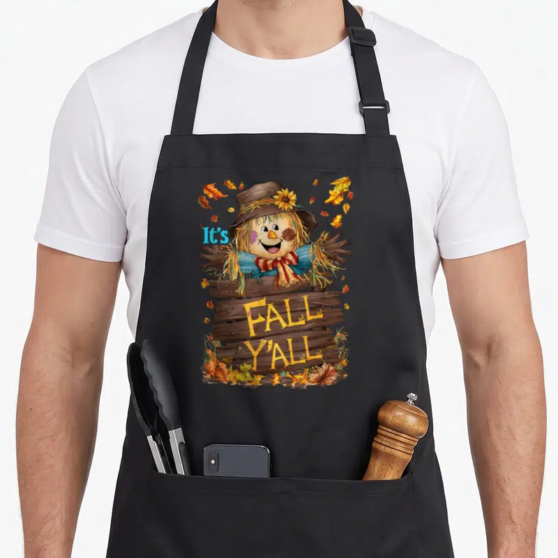 Fall yall thanksgiving apron - Cartoon scarecrow behind wooden sign reading It's FALL Y'ALL surrounded by brown yellow — shown on a black cotton canvas apron against a white background