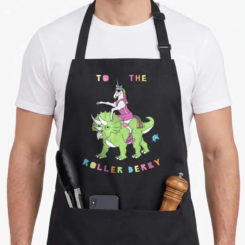 Roller dinosaur apron - Cartoon unicorn in pink skates riding a green triceratops with multi-colored TO THE ROLLER — shown on a black cotton canvas apron against a white background