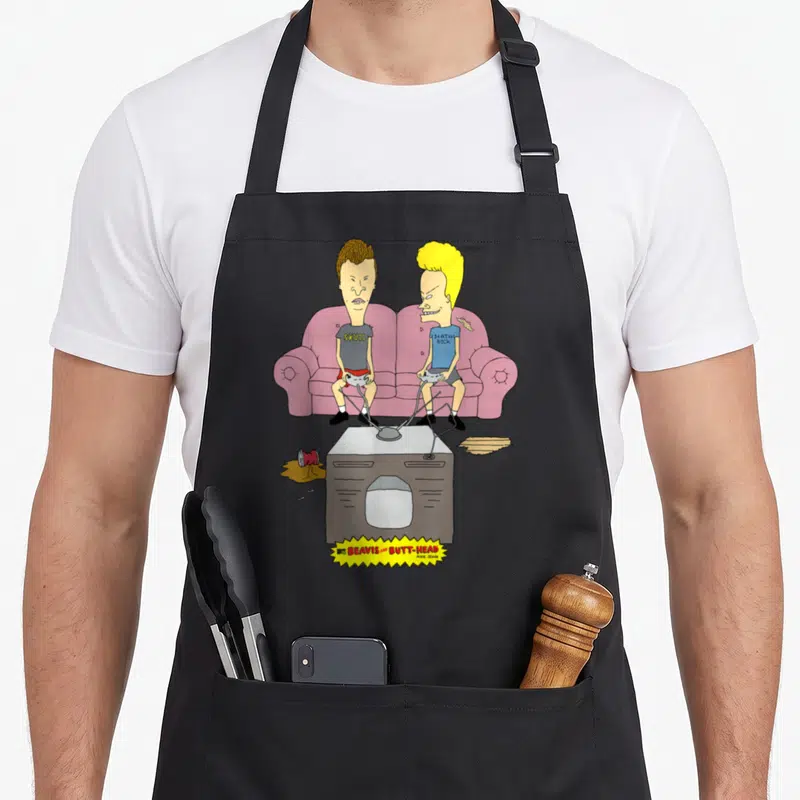 Beavis game aprons - Two cartoon men on a pink sofa holding controllers with skull and death rock text below — shown on a black cotton canvas apron against a white background