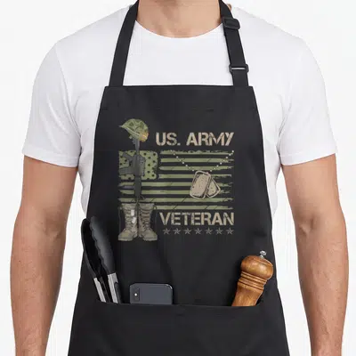 Army patriotic apron - US Army Veteran text with helmet rifle boots and dog tags on faded flag — shown on a black cotton canvas apron against a white background