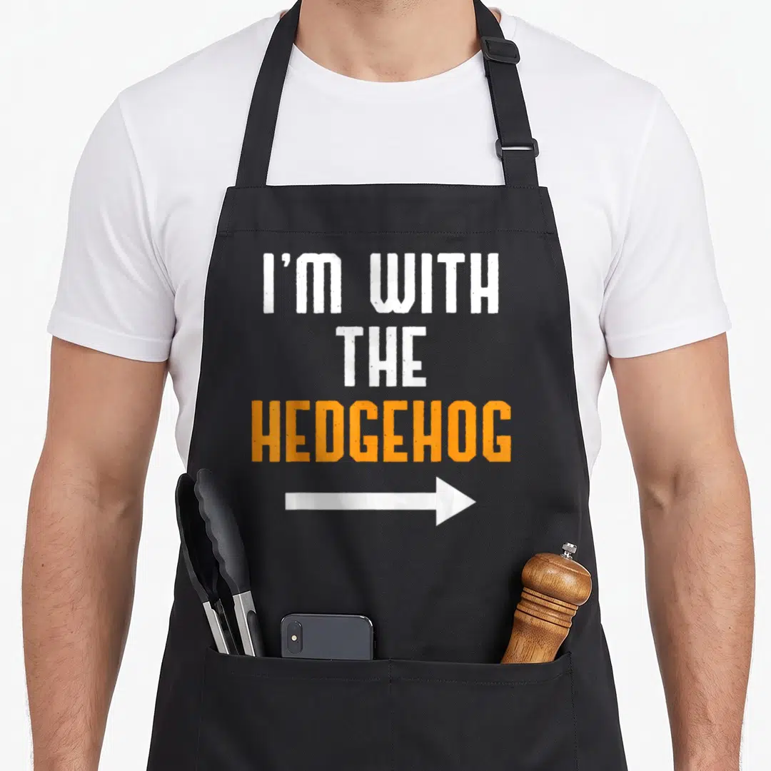 Hedgehog couple apron - Stacked white and orange text reading I'm With The Hedgehog above a white right-pointing arrow — shown on a black cotton canvas apron against a white background