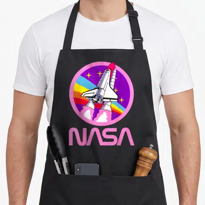 Nerdy nasa apron - Cartoon space shuttle in a pink circle with rainbow stripes and large pink NASA text below — shown on a black cotton canvas apron against a white background