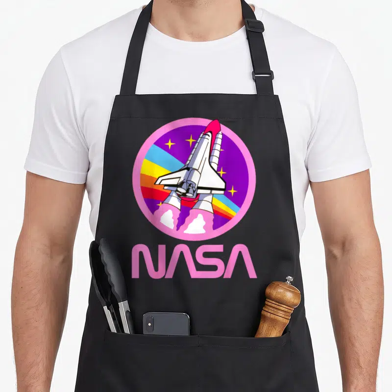 Nerdy nasa apron - Cartoon space shuttle in a pink circle with rainbow stripes and large pink NASA text below — shown on a black cotton canvas apron against a white background