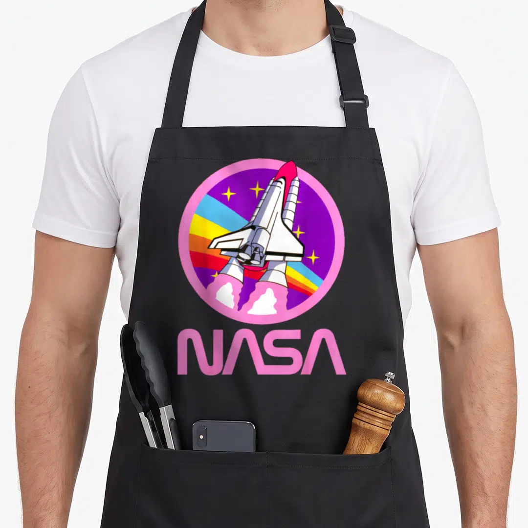 Nerdy nasa apron - Cartoon space shuttle in a pink circle with rainbow stripes and large pink NASA text below — shown on a black cotton canvas apron against a white background