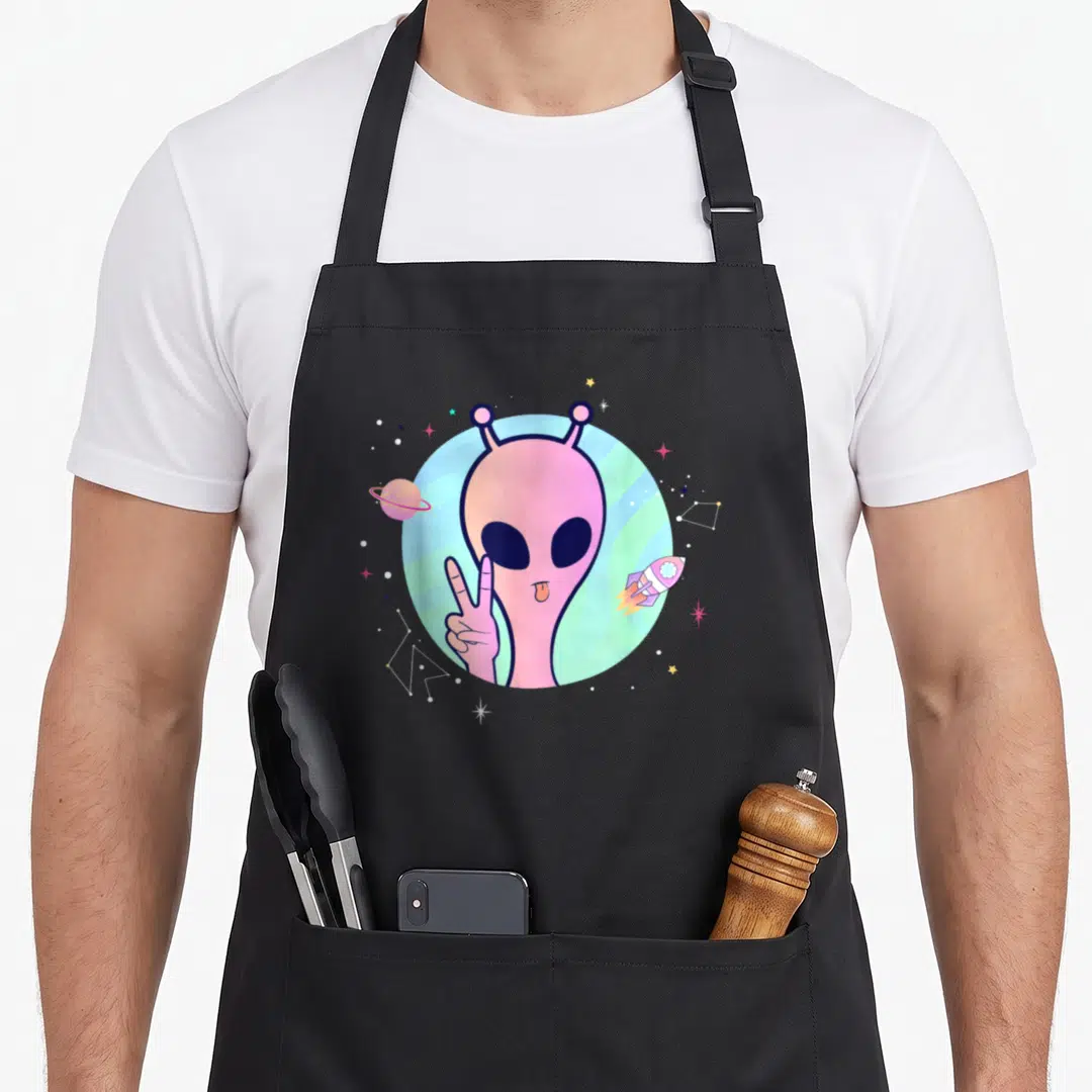 Peace ping apron - Cartoon pink alien making a peace sign with planet, rocket, and stars on blue-green gradient — shown on a black cotton canvas apron against a white background