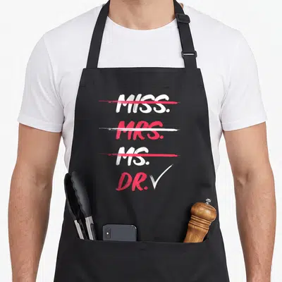 Miss dr apron - White and red text listing Miss Mrs Ms crossed out and Dr checked vertically — shown on a black cotton canvas apron against a white background
