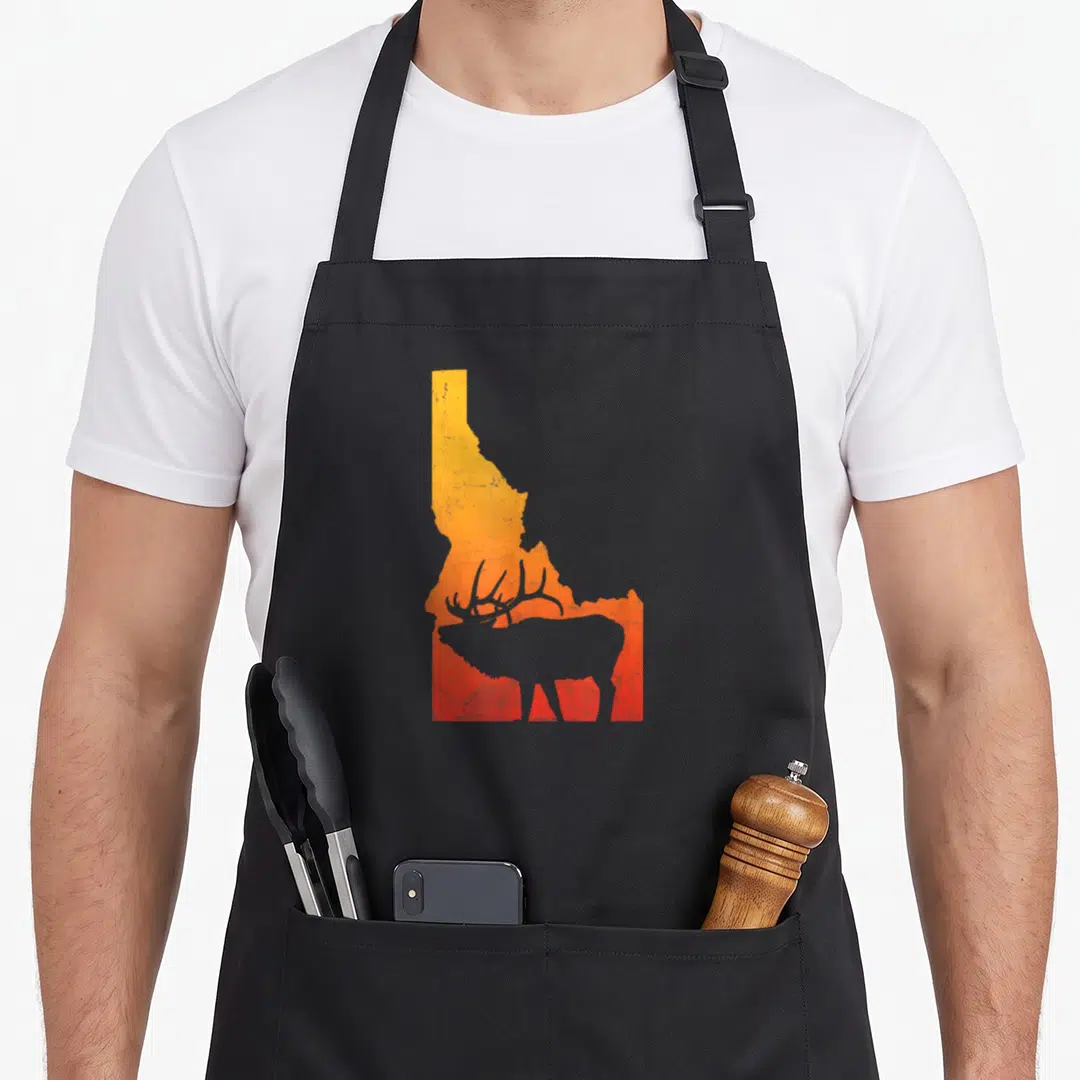 Idaho elk outdoor apron - Orange to red gradient Idaho state map with grey elk silhouette cutout in lower section — shown on a black cotton canvas apron against a white background