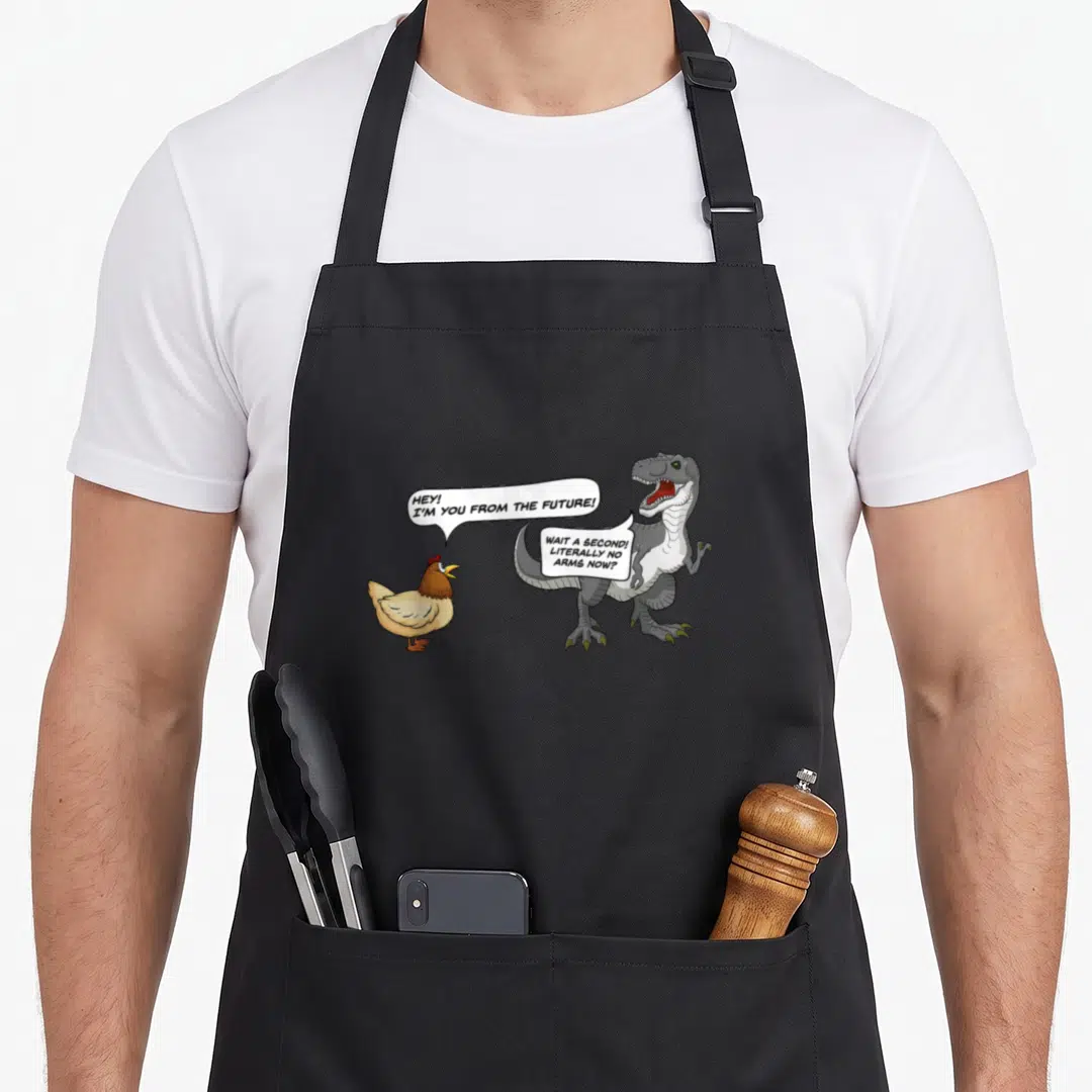 Chicken t rex apron - Cartoon chicken and T-Rex with speech bubbles about evolution and missing arms — shown on a black cotton canvas apron against a white background