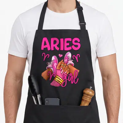 Sneaker aries kitchen apron - ARIES girl text with pink sneakers, gold rings, and zodiac ram symbols in cartoon art — shown on a black cotton canvas apron against a white background