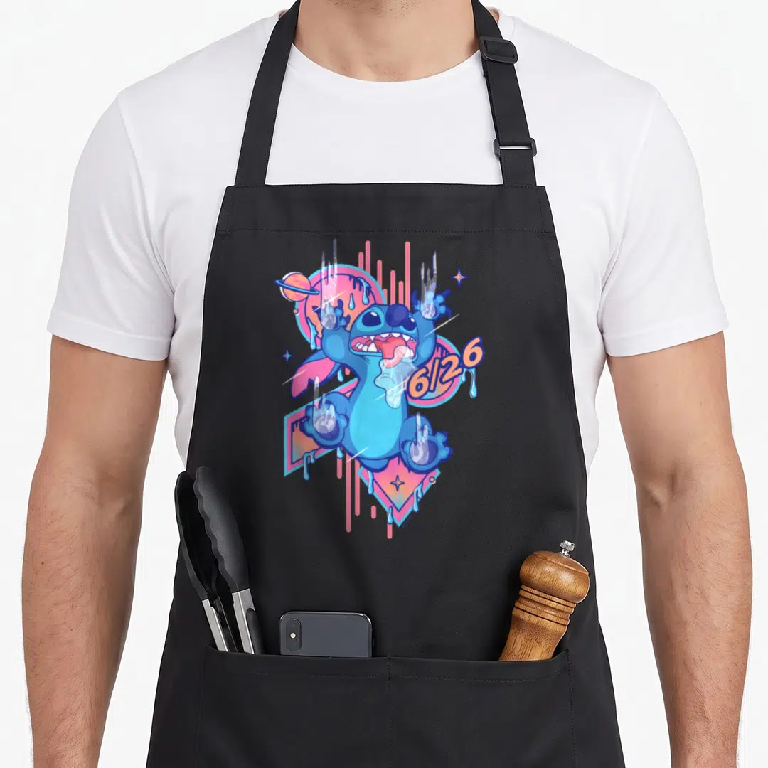 Drool stitch apron - Blue cartoon figure with 6/26 text, pink circle, orange drips, and hand prints — shown on a black cotton canvas apron against a white background