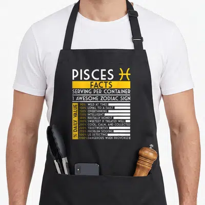 Pisce fact zodiac aprons - Nutrition facts style graphic listing Pisces traits like loyal and intelligent with yellow — shown on a black cotton canvas apron against a white background