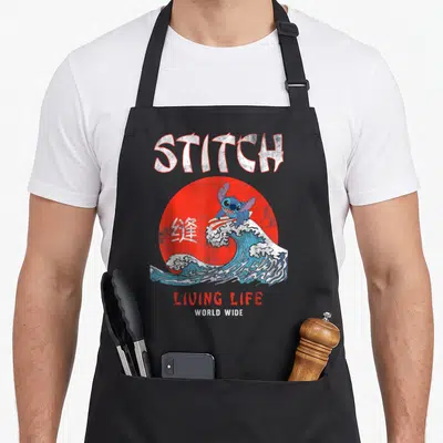 Living stitch apron - Blue character surfing on wave in front of red sun with STITCH and Chinese text — shown on a black cotton canvas apron against a white background