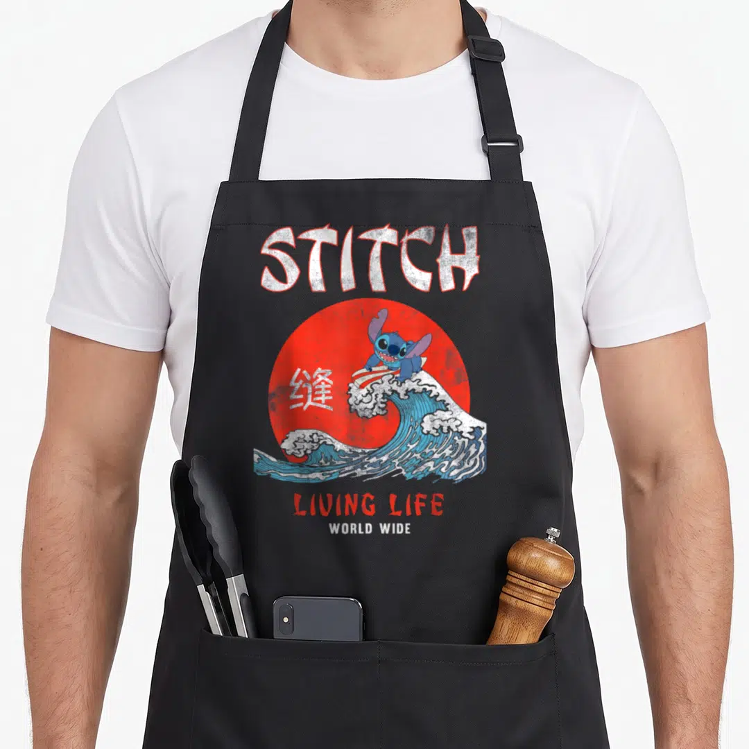 Living stitch apron - Blue character surfing on wave in front of red sun with STITCH and Chinese text — shown on a black cotton canvas apron against a white background