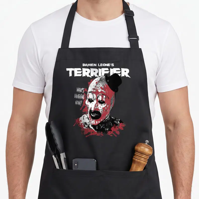 Distressed clown aprons - Distressed clown head with red splatters and text reading Damien Leone's Terrifier Who's — shown on a black cotton canvas apron against a white background