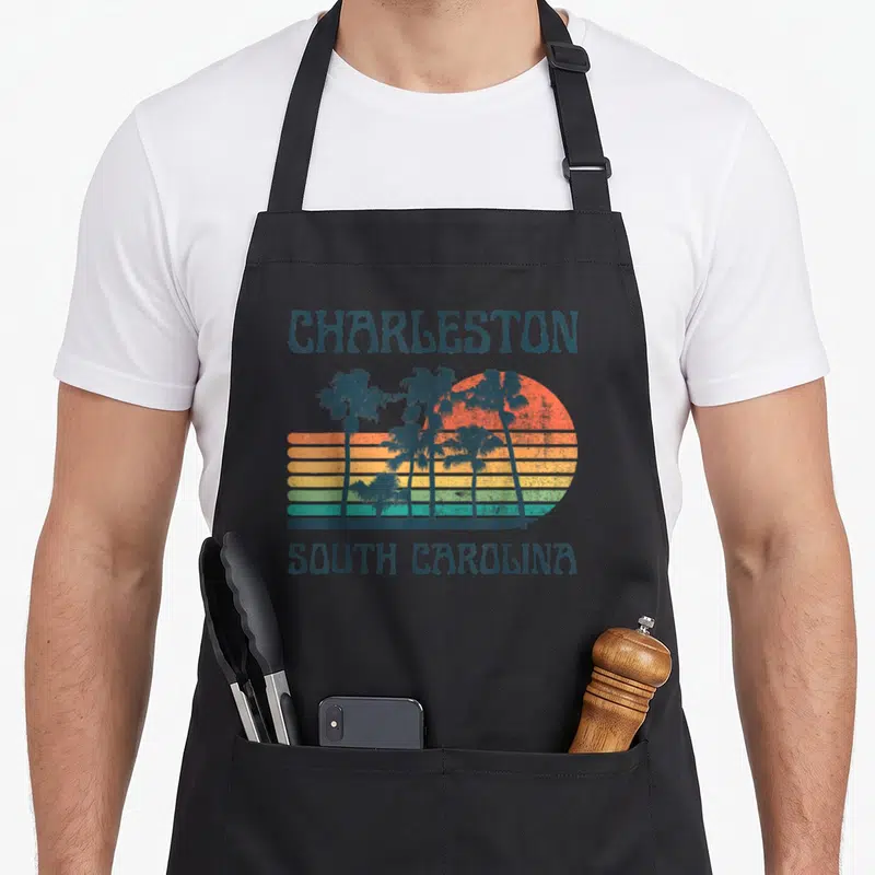 Charleston summer aprons - Retro sunset with palm trees and Charleston South Carolina text in blue grey orange yellow — shown on a black cotton canvas apron against a white background