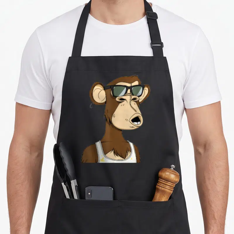 Bored ape apron - Cartoon brown monkey wearing black sunglasses and white tank top with yellow stains — shown on a black cotton canvas apron against a white background