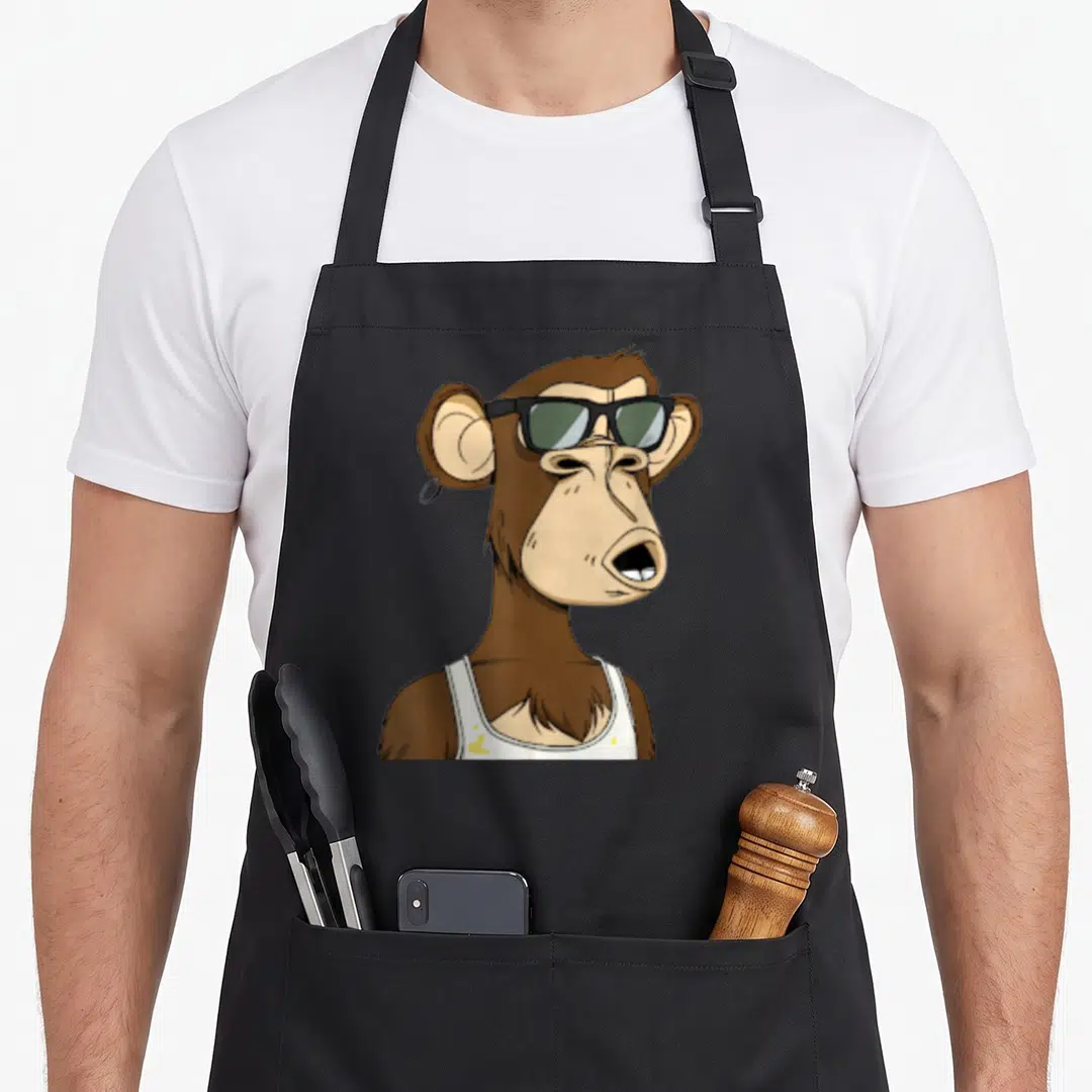 Bored ape apron - Cartoon brown monkey wearing black sunglasses and white tank top with yellow stains — shown on a black cotton canvas apron against a white background