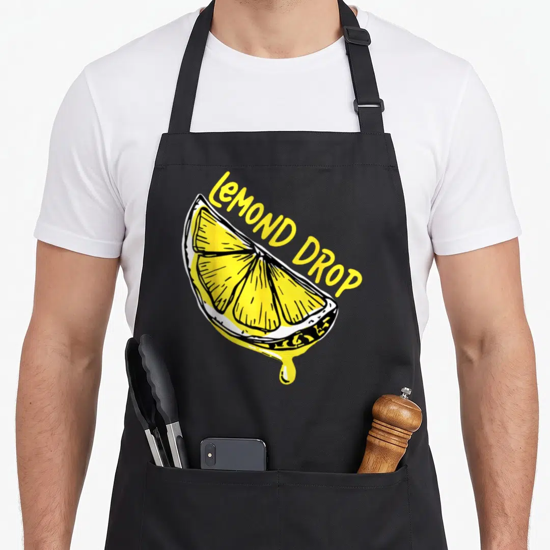 Lemondrop for love and lemons apron - Yellow cartoon lemon slice with arched LEMOND DROP text and a single dripping — shown on a black cotton canvas apron against a white background
