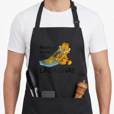 Lasagna cat apron - Orange cartoon cat eating from blue pan with Mmmmmm text and LASAGNA label — shown on a black cotton canvas apron against a white background