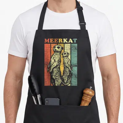 Cool meerkat apron - Two meerkats standing in front of red, orange, yellow, and green vertical stripes under the word — shown on a black cotton canvas apron against a white background