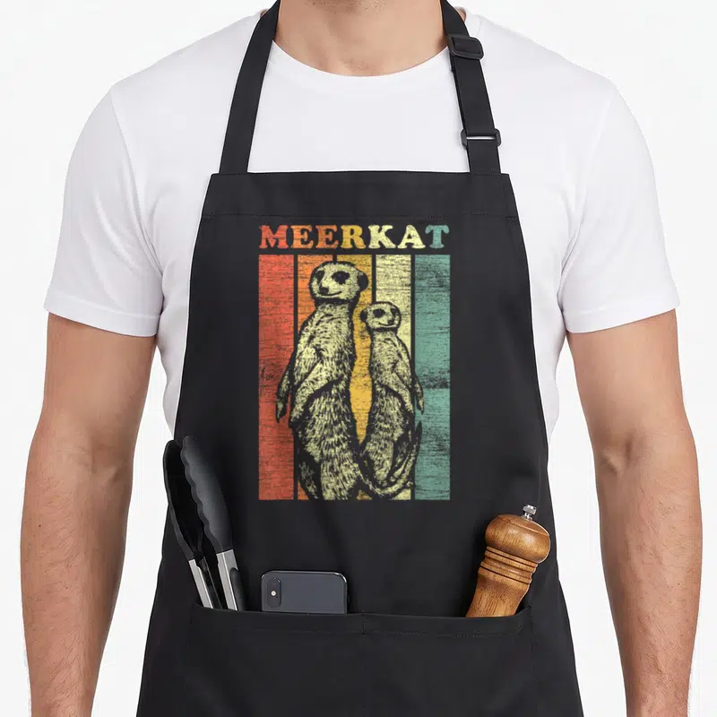 Cool meerkat apron - Two meerkats standing in front of red, orange, yellow, and green vertical stripes under the word — shown on a black cotton canvas apron against a white background