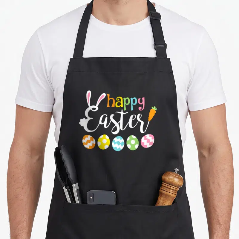 Rabbit face easter bunny aprons - Happy Easter text with bunny ears, carrot, and five patterned eggs in pastel colors — shown on a black cotton canvas apron against a white background