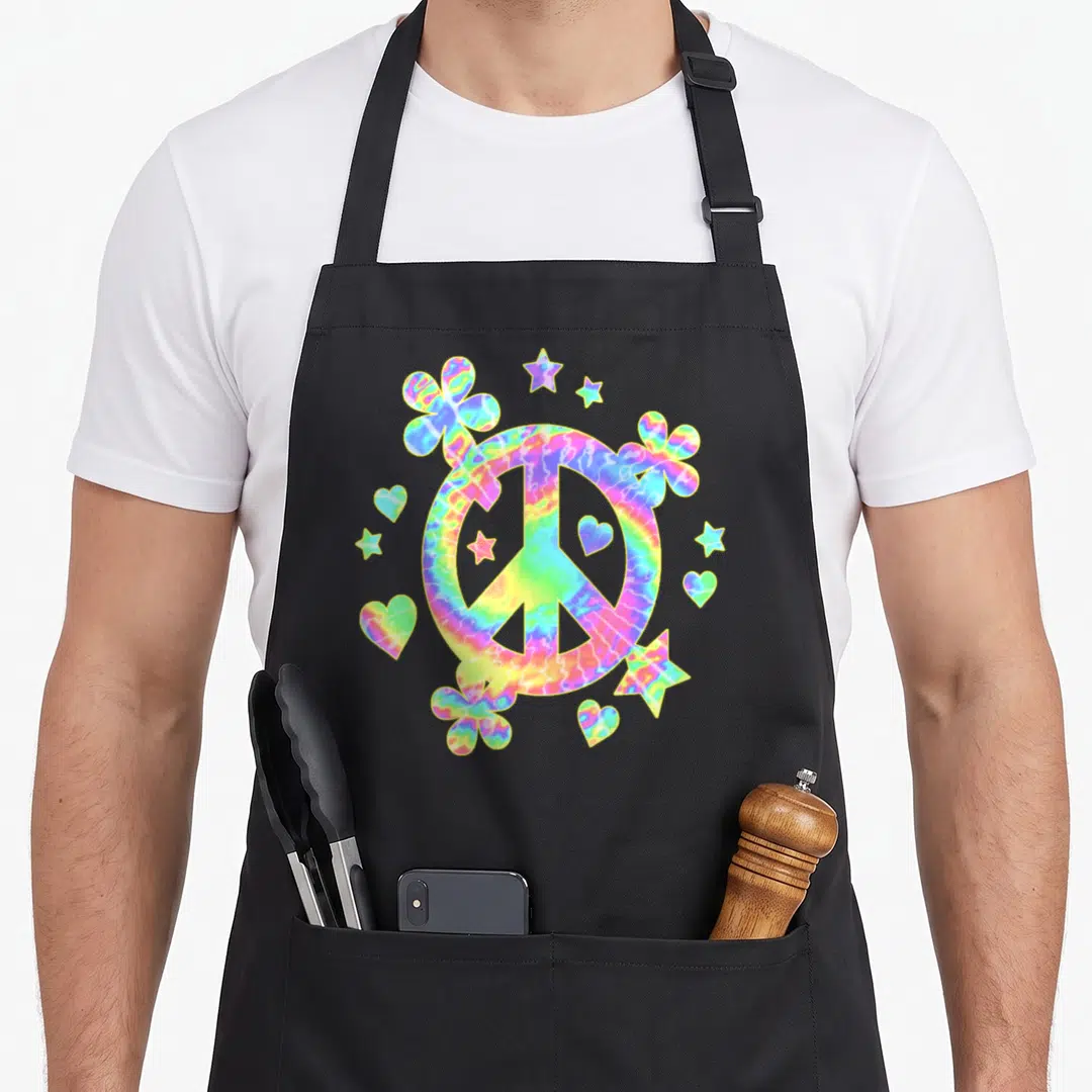 Sign tie dye apron - Colorful tie-dye peace sign surrounded by flowers, hearts, and stars in pink, yellow, blue — shown on a black cotton canvas apron against a white background