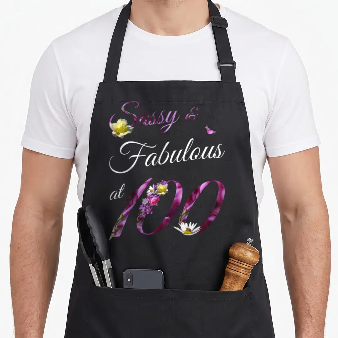 Sassy floral aprons - Stacked purple and white text reading Sassy & Fabulous at 100 with floral overlays — shown on a black cotton canvas apron against a white background