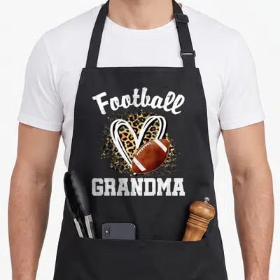 Leopard grandma aprons - Football GRANDMA text above leopard heart and brown football graphic — shown on a black cotton canvas apron against a white background