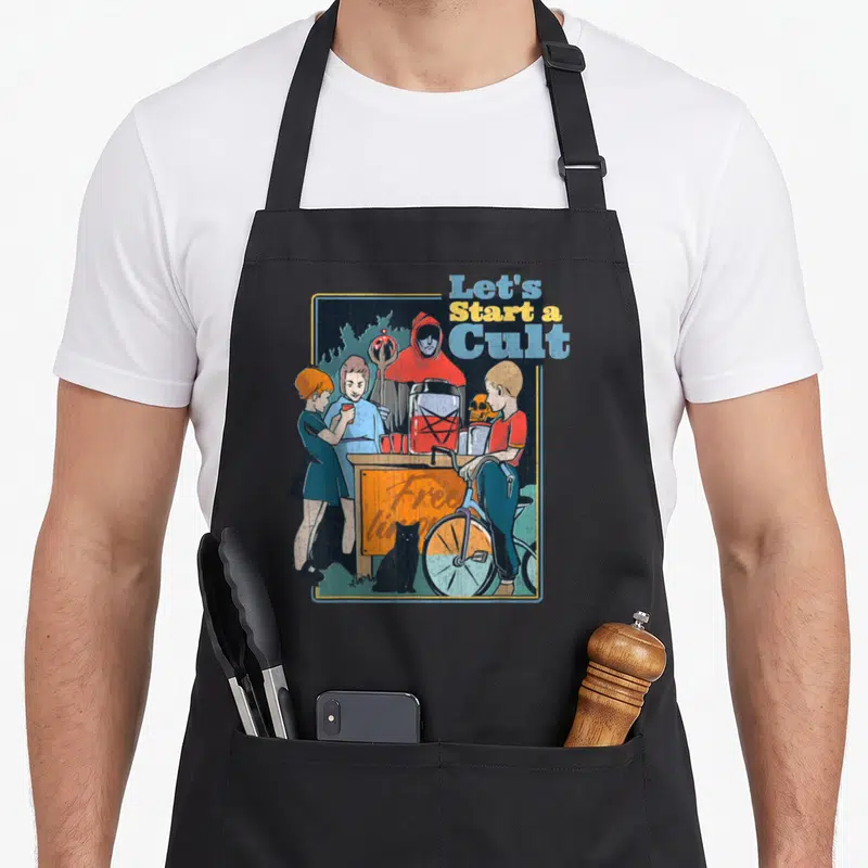 Start game aprons - Hooded figure behind table with children and cat, text Let's Start a Cult in red blue yellow grey — shown on a black cotton canvas apron against a white background
