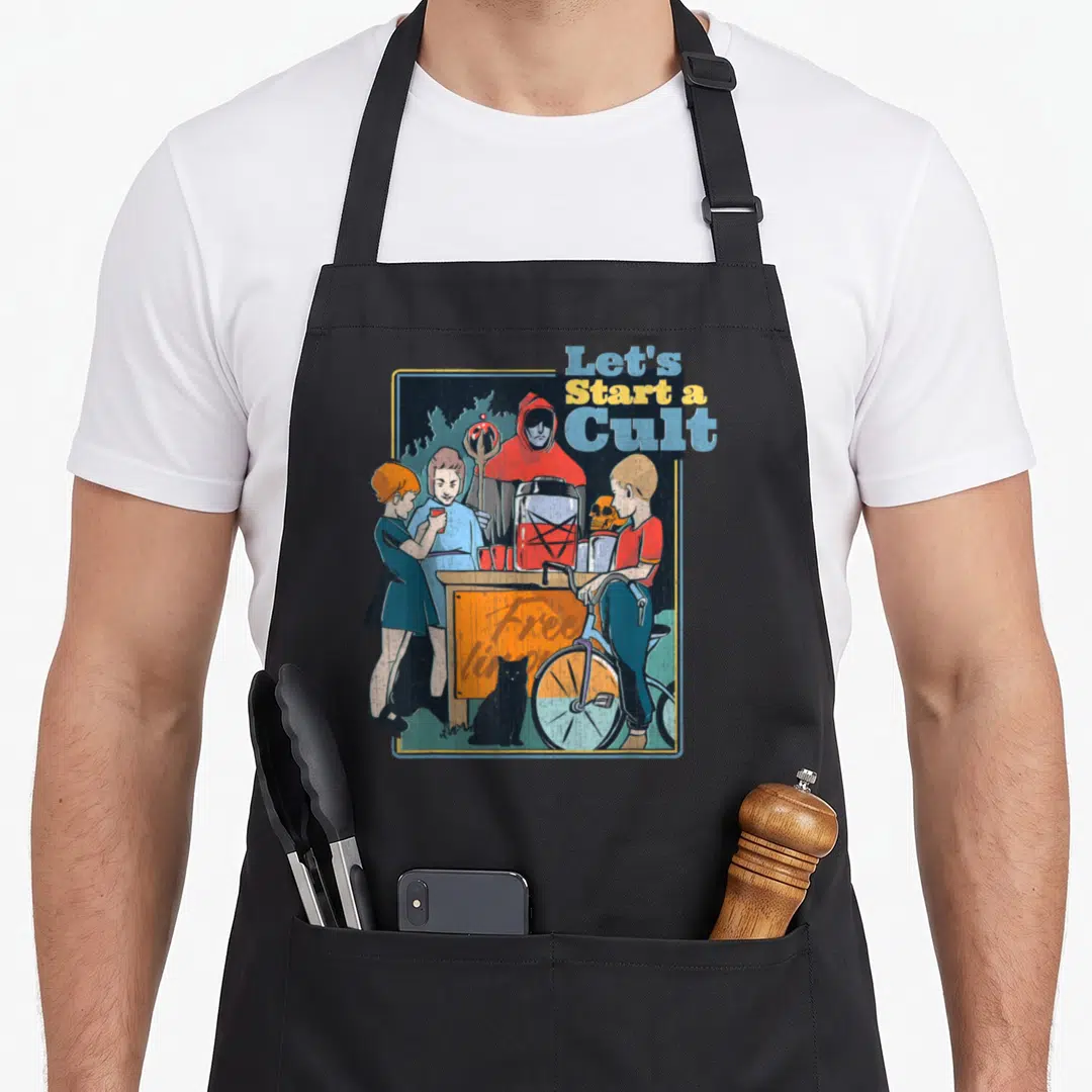 Start game aprons - Hooded figure behind table with children and cat, text Let's Start a Cult in red blue yellow grey — shown on a black cotton canvas apron against a white background