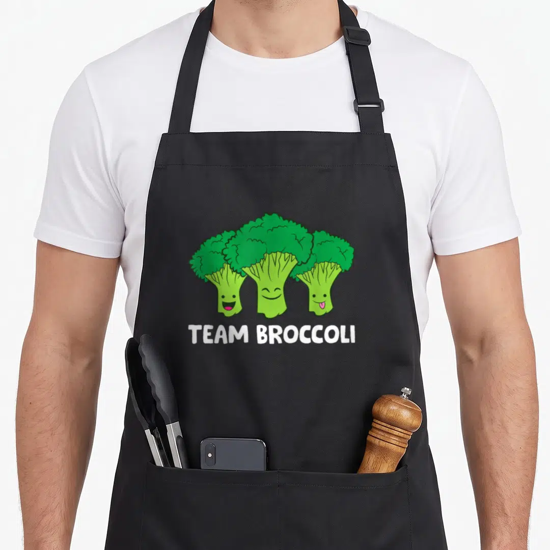 Broccoli vegan apron - Three cartoon broccoli characters with smiling faces and white TEAM BROCCOLI text — shown on a black cotton canvas apron against a white background