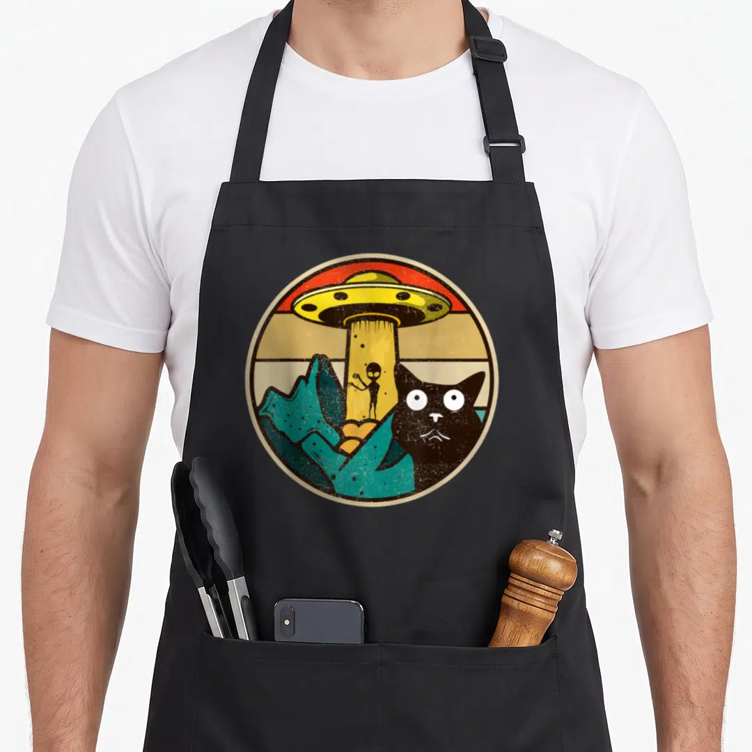 Ufo black cat aprons - Circular cartoon of black cat, yellow UFO beam, teal mountains, and red orange beige stripes — shown on a black cotton canvas apron against a white background
