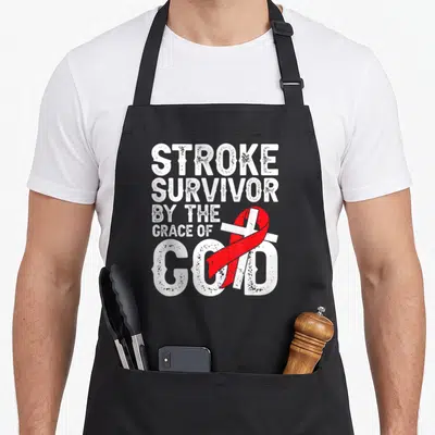 Stroke grace apron - Stacked white text Stroke Survivor By The Grace Of God with distressed cross and red ribbon — shown on a black cotton canvas apron against a white background
