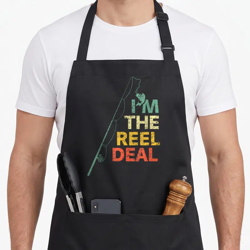 Deal fisherman apron - Fishing rod diagonal left, four stacked words I'M THE REEL DEAL in green beige yellow red retro — shown on a black cotton canvas apron against a white background
