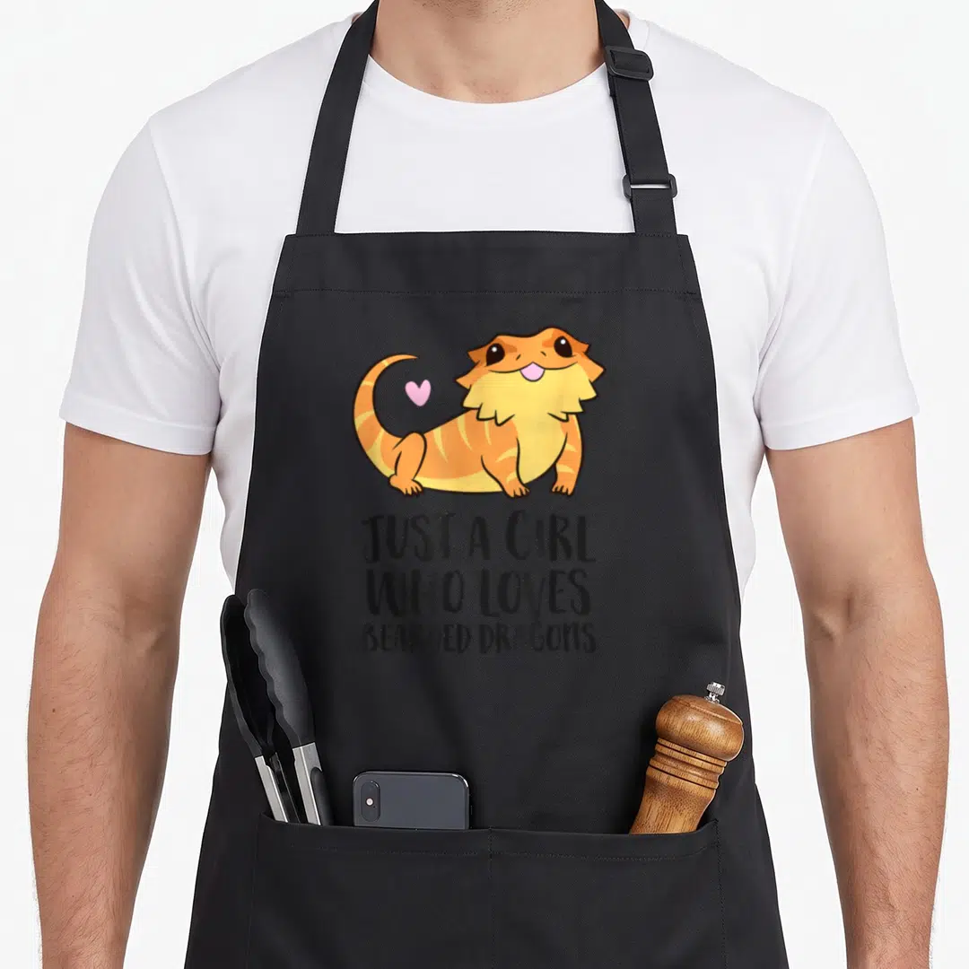 Vintage bearded dragon apron - Cartoon orange bearded dragon with pink heart above stacked black text reading Just — shown on a black cotton canvas apron against a white background