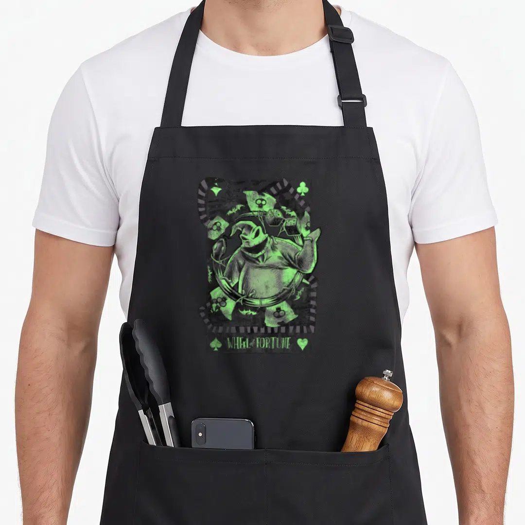 Card christmas apron - Neon green monster surrounded by dice, ribbons, skull, and WHEEL of FORTUNE text with card suit — shown on a black cotton canvas apron against a white background