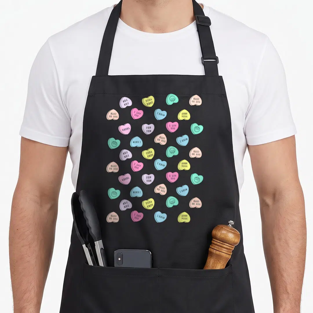 Valentine cotton candy apron - Scattered pink yellow blue and green candy hearts with Star Wars puns like Yoda Best — shown on a black cotton canvas apron against a white background