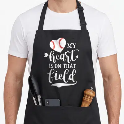 Field softball baseball apron - White heart with red stitching and arrow through text MY heart IS ON THAT field — shown on a black cotton canvas apron against a white background