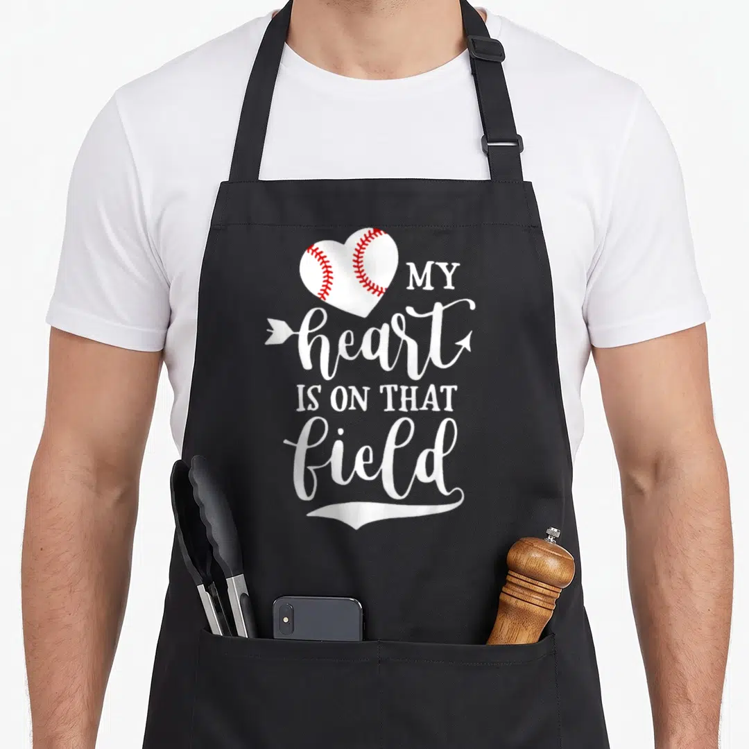 Field softball baseball apron - White heart with red stitching and arrow through text MY heart IS ON THAT field — shown on a black cotton canvas apron against a white background
