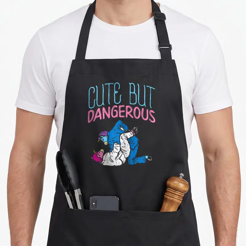 Boredkoalas cartoon apron - CUTE BUT DANGEROUS text above two wrestling unicorns in blue and pink — shown on a black cotton canvas apron against a white background