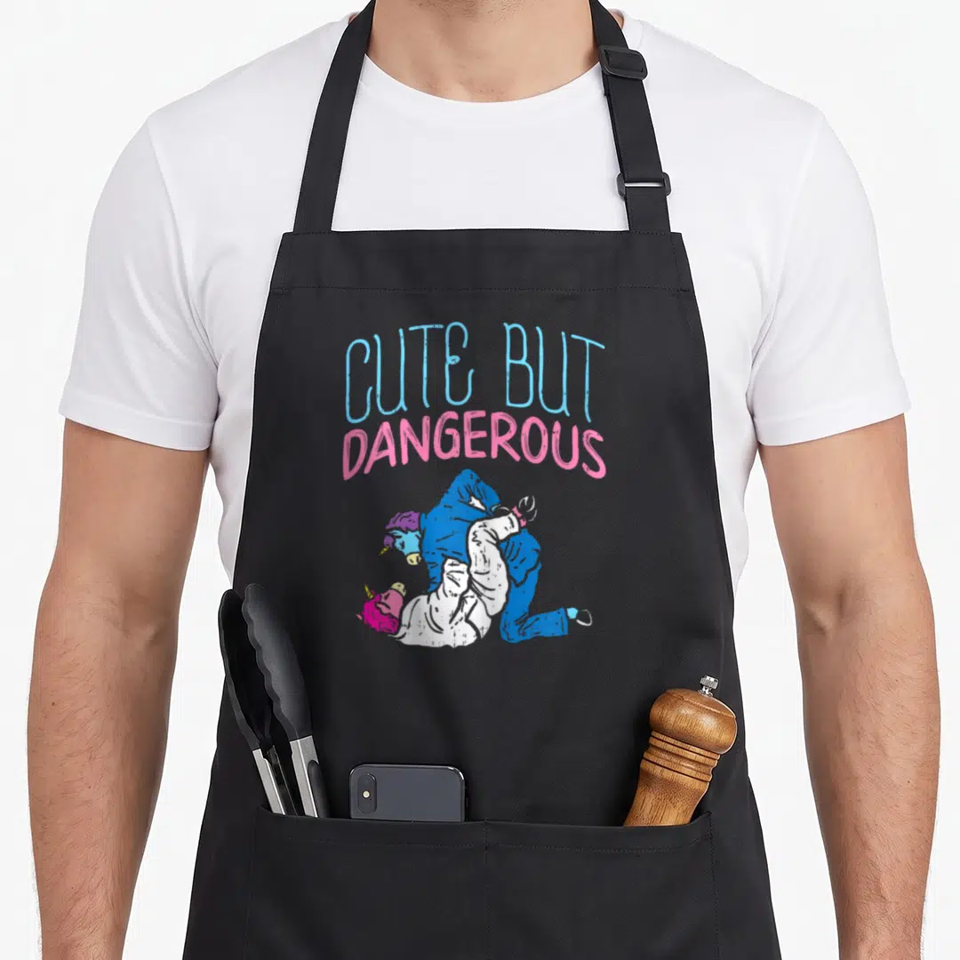 Boredkoalas cartoon apron - CUTE BUT DANGEROUS text above two wrestling unicorns in blue and pink — shown on a black cotton canvas apron against a white background