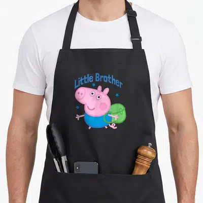 Brother pig apron - Pink pig in blue shirt and green backpack under arched Little Brother text with blue stars — shown on a black cotton canvas apron against a white background